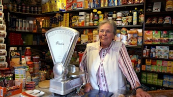 Eine ältere Dame mit Brille steht hinter einer Ladentheke, hinter ihr sind Regale mit Kaffee, Tee und anderen Lebensmitteln zu sehen, eine altmodische Waage steht neben.
