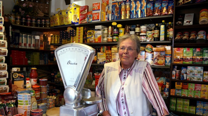 Eine ältere Dame mit Brille steht hinter einer Ladentheke, hinter ihr sind Regale mit Kaffee, Tee und anderen Lebensmitteln zu sehen, eine altmodische Waage steht neben.&nbsp;
