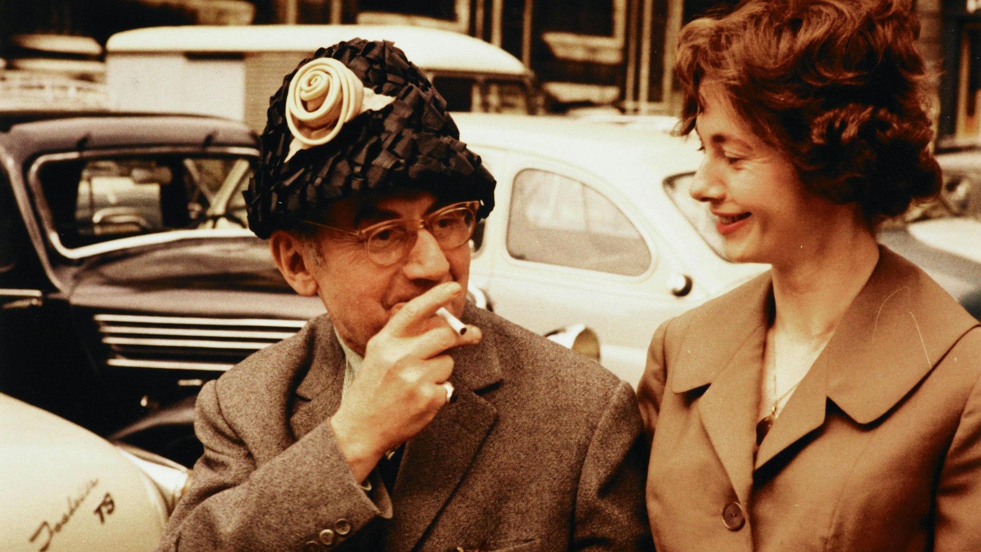 Renate Gruber schaut lächelnd auf den rauchenden Künstler Man Ray, der zum Spaß ihren Hut trägt.