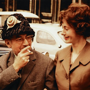 Renate Gruber schaut lächelnd auf den rauchenden Künstler Man Ray, der zum Spaß ihren Hut trägt.