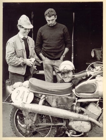 Auf einem Schwarz-Weiß-Bild ist eine junge Frau mit Käppi zu sehen, vor ihr kniet ein Mann mit Helm hinter einem Motorrad mit der Startnummer 247.