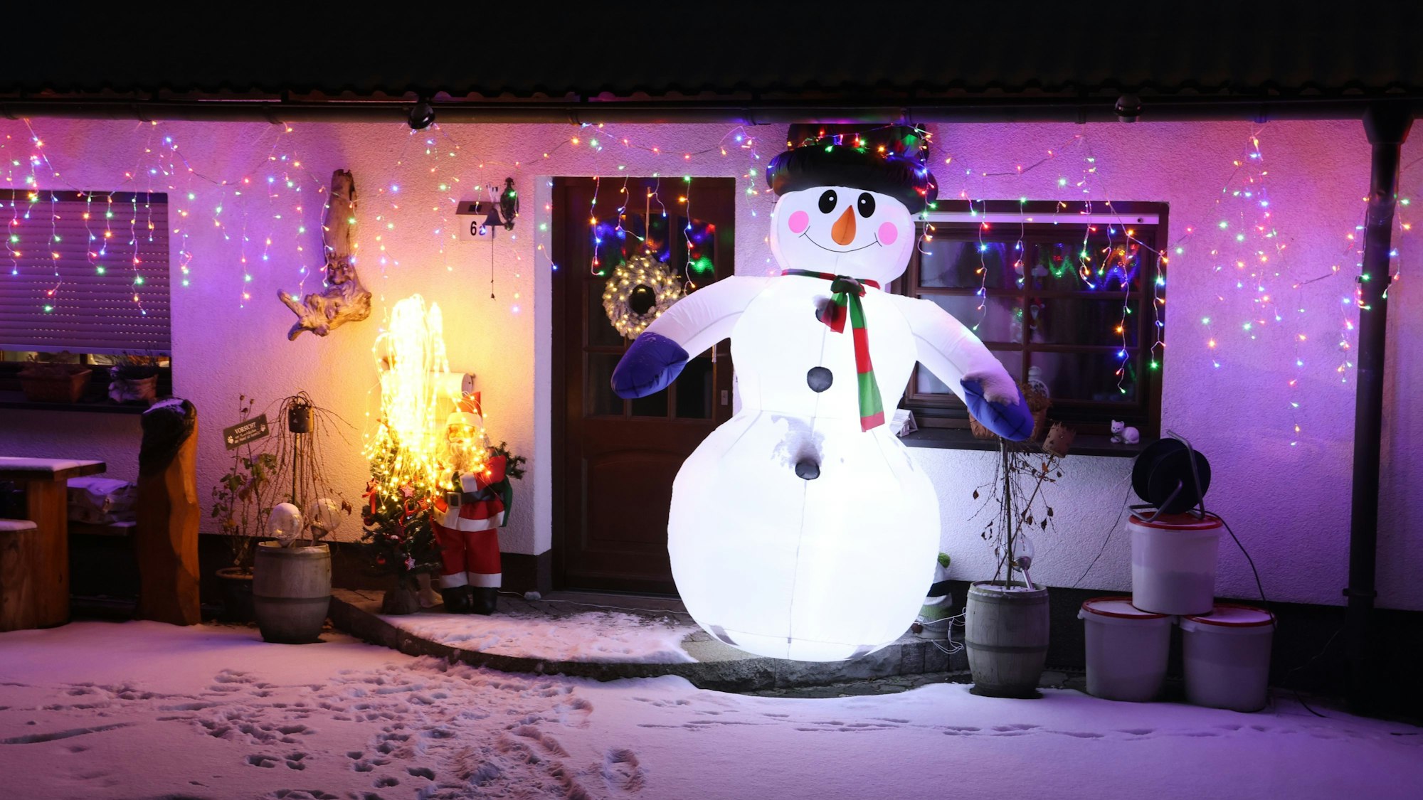 Ein großer aufblasbarer Schneemann steht vor einer Haustüre. Im Hintergrund hängen bunte Lichterketten an der Dachrinne des Hauses.