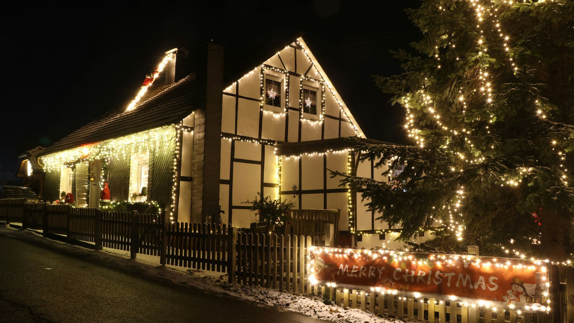 Die Konturen eines alten Fachwerkhauses sind mit Lichterketten nachgezeichnet. Am Zaun hängt ein rotes Banner, auf dem "Merry Christmas“ steht.