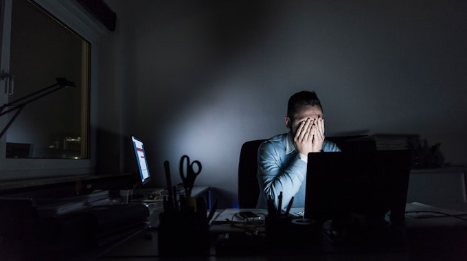 Ein Mann sitzt in einem dunklen Büro am Computer und hält sich die Hände vors Gesicht.
