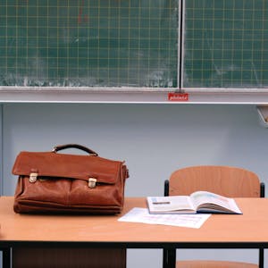 Auf einem Pult liegt eine braune Lehrertasche. Im Hintergrund ist eine leere Tafel zu sehen.