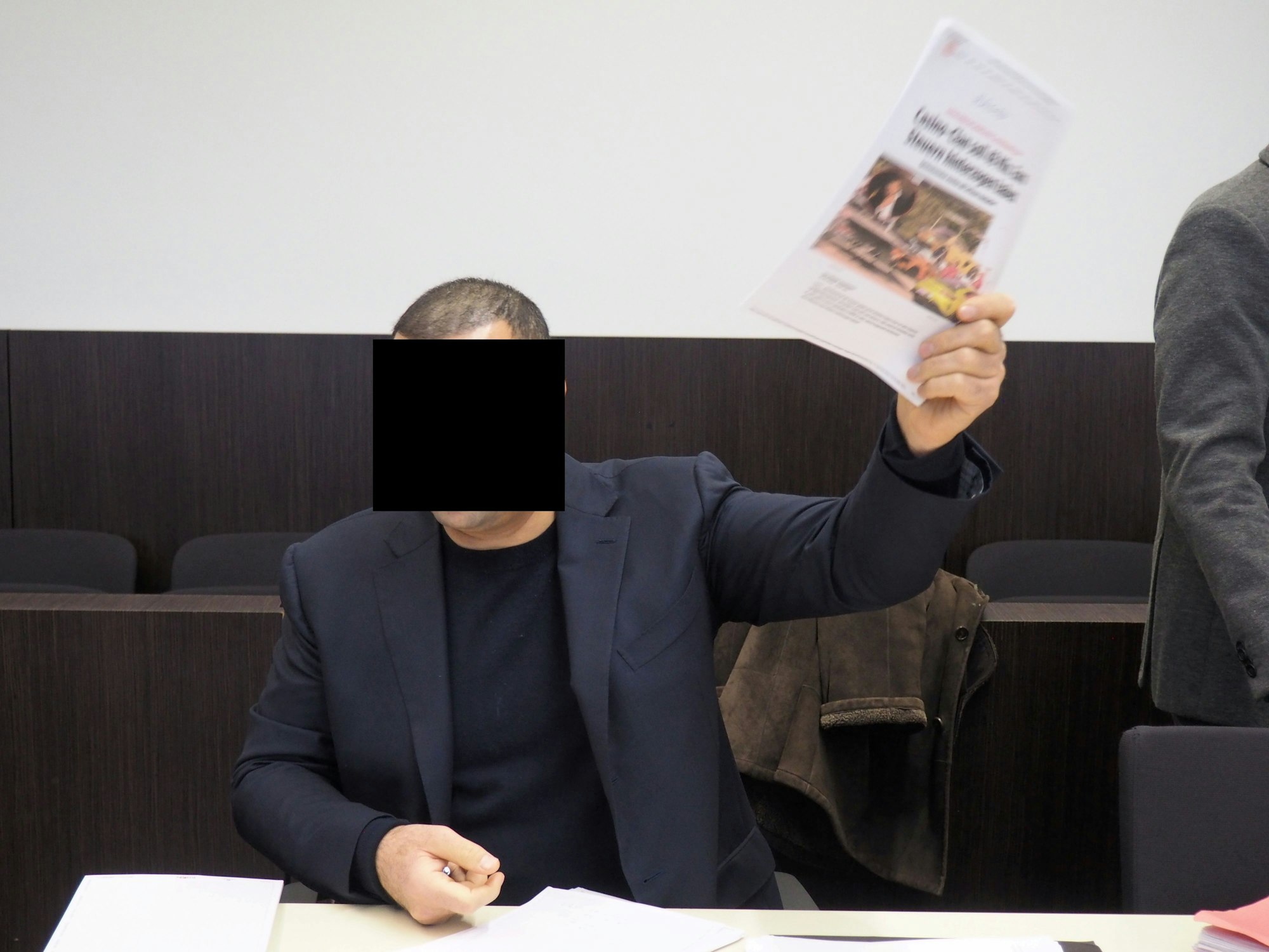 Mehmet R. sitzt im Anzug im Gerichtsaal und hält einen Zeitungsartikel hoch, in dem über die Festnahme eines Konkurrenten berichtet wurde.