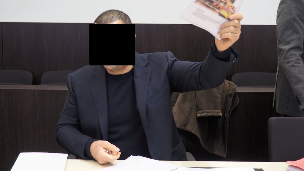 Mehmet R. sitzt im Anzug im Gerichtsaal und hält einen Zeitungsartikel hoch, in dem über die Festnahme eines Konkurrenten berichtet wurde.