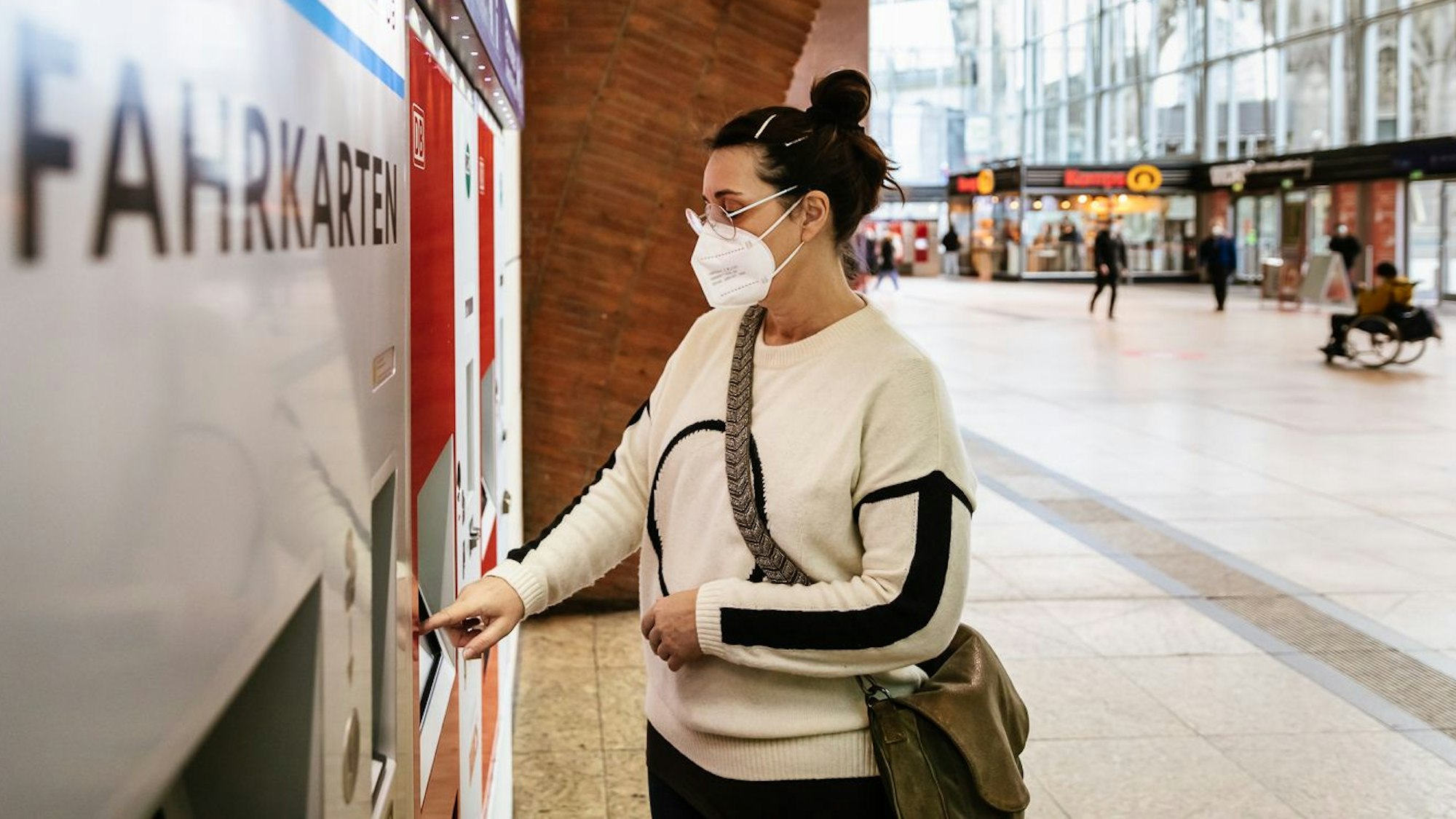 Eine Frau mit Maske zieht sich im Kölner Hauptbahnhof ein Ticket aus einem VRS-Automaten.