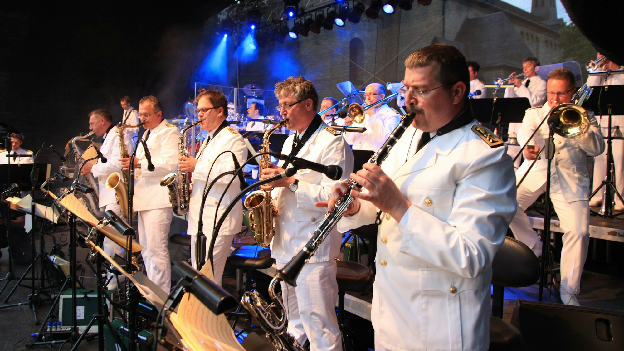 Musiker der Bundeswehr Big Band stehen auf einer Bühne in blauem Scheinwerferlicht, sie tragen weiße Uniformen. Sie spielen Saxophon, Trompete und Klarinette.