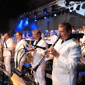 Musiker der Bundeswehr Big Band stehen auf einer Bühne in blauem Scheinwerferlicht, sie tragen weiße Uniformen. Sie spielen Saxophon, Trompete und Klarinette.