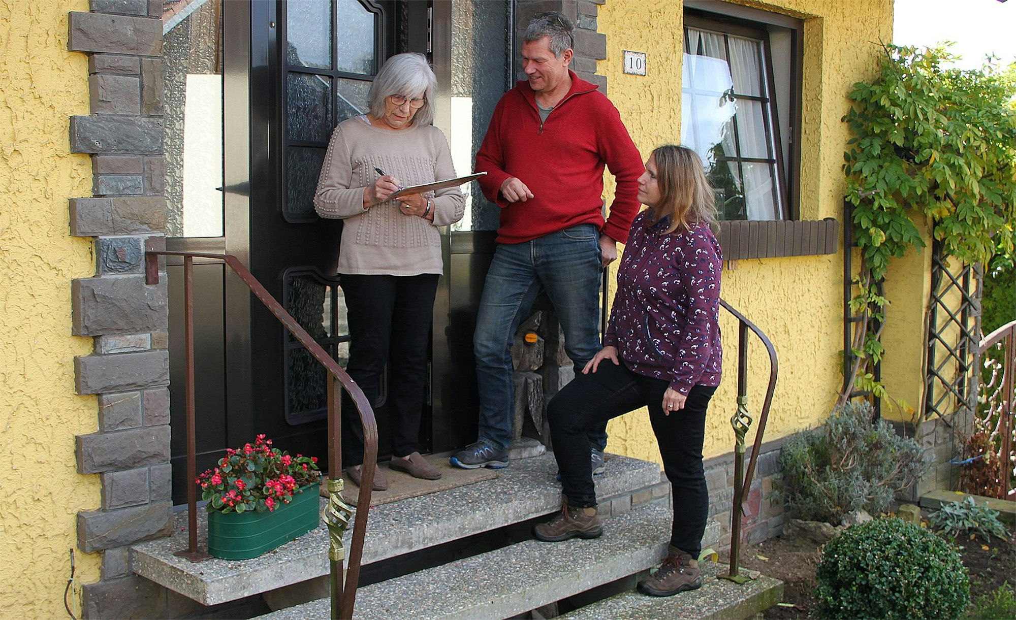 Drei Personen stehen im Eingang eines gelben Hauses, eine Frau unterschreibt auf einem Klemmbrett.