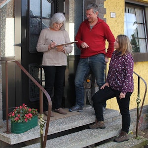 Drei Personen stehen im Eingang eines gelben Hauses, eine Frau unterschreibt auf einem Klemmbrett.