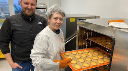 Kerstin Hottewitzsch und Nicolas Gritto stehen vor einem Industrie-Ofen. Hottewitzsch hält ein Blech mit Keksen und schiebt es in den Ofen. Sie lächeln beide in die Kamera.