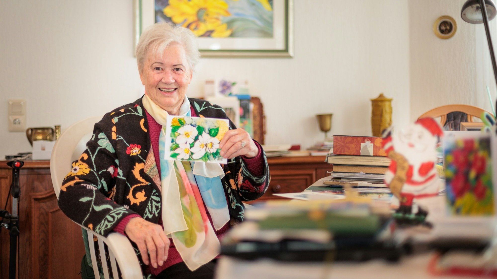 Marie-Luise Stricker hält eine bunte und selbstgemalte Weihnachtskarte in der Hand. Auf dem Tisch vor ihre liegen zahlreiche weitere Karten.