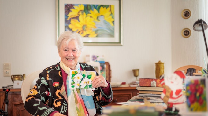 Marie-Luise Stricker hält eine bunte und selbstgemalte Weihnachtskarte in der Hand. Auf dem Tisch vor ihre liegen zahlreiche weitere Karten.