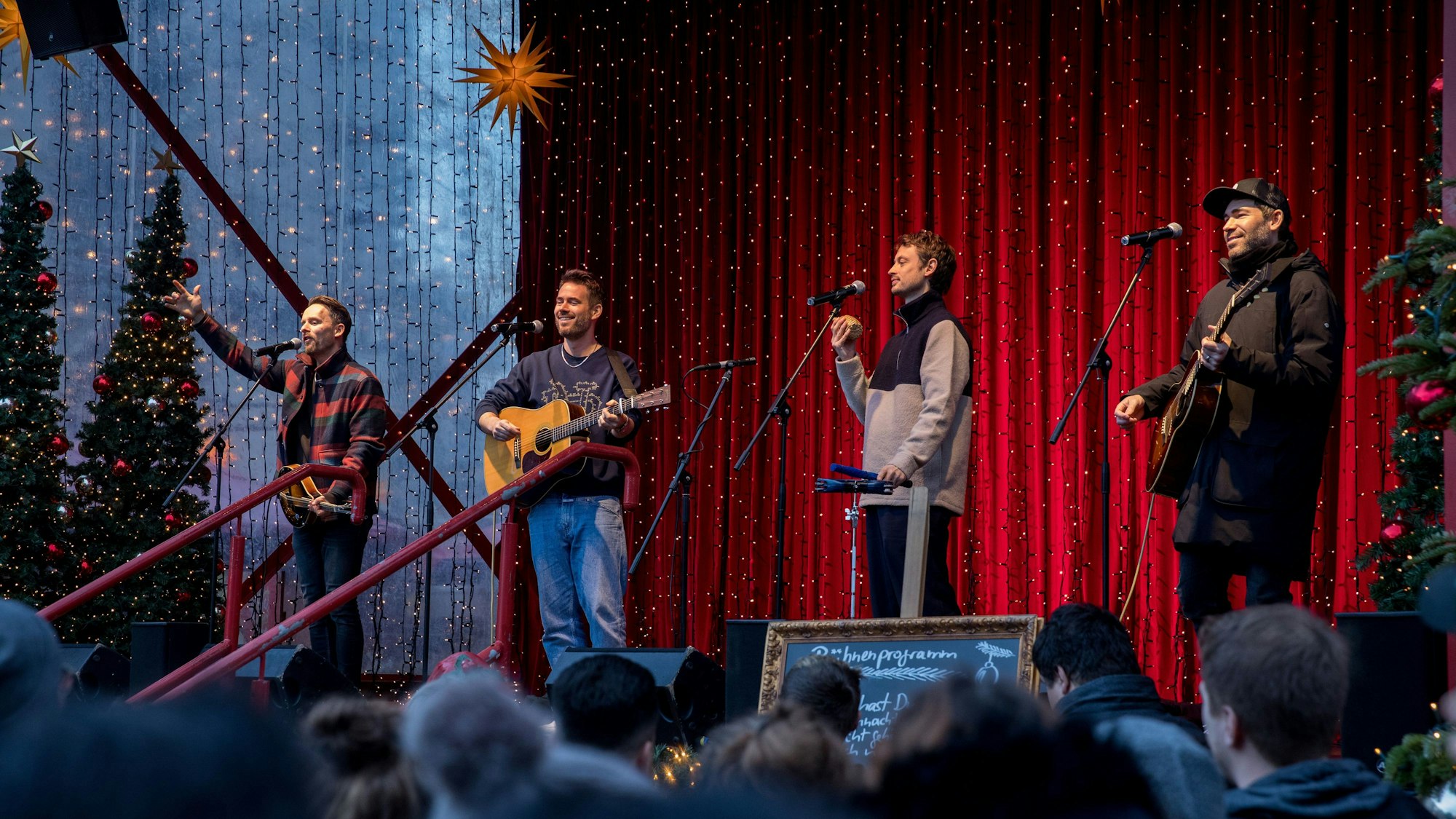 Livekonzert der Band Revolverheld auf dem Weihnachtsmarkt am Dom. Die vier Musiker an Mikros vor leuchtenden Lichterketten und rotem Vorhang.