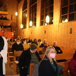 Ein grauhaariger Mann mit Schnauzer steht im weißen Messgewand und einem schwarzen Schal in einer Kirche inmitten von halb besetzten Kirchenbänken.