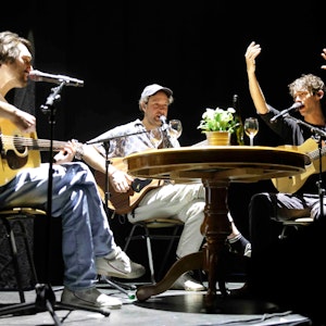 Die Band AnnenMayKantereit sitzt auf der Bühne der Lanxess-Arena an einem Tisch und performt.