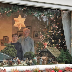 Ein älteres Paar steht hinter dem Fenster. Im Schaufenster sieht man eine handgemachte Krippe.