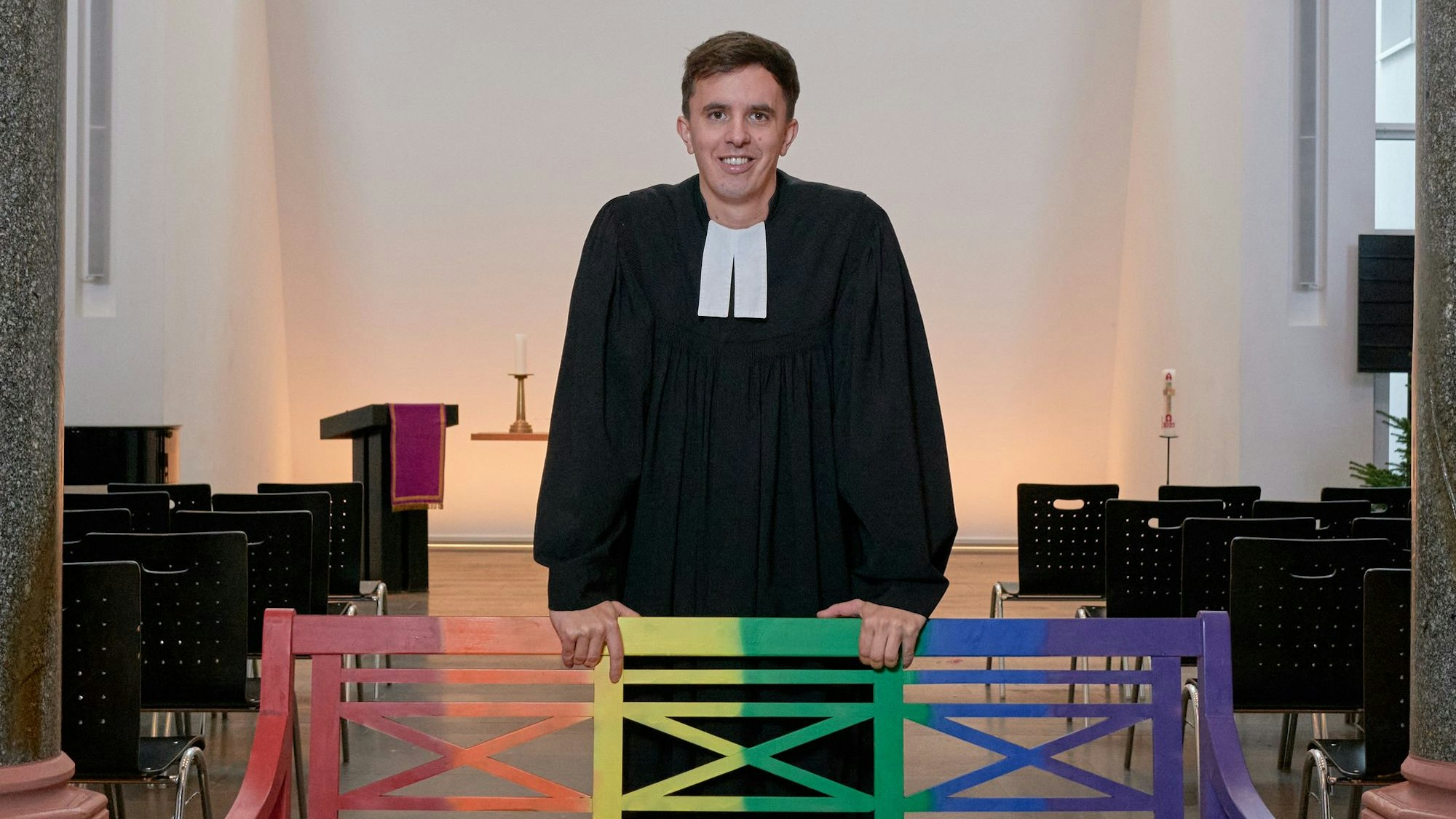 Tim Lahr, evangelischer Pfarrer, steht im Talar in der Kölner Christuskirche am Stadtgarten. Er stützt sich mit den Händen auf eine Holzbank, die in Regenbogenfarben gestrichen ist.