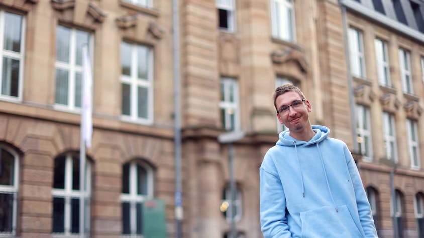 Kommunikative Kompetenz, Ideen und Erfahrung bringt Florian Lintz (32) in seinen Lehrauftrag ein – unter anderem. Er steht entspannt in einem hellblauen Sweatshirt vor dem Lehrgebäude am Ubierring.