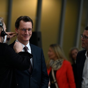 Ein Tontechniker befestigt ein Mikrofon am Gesicht von NRW-Ministerpräsident Hendrik Wüst. Der lächelt in die Kamera.