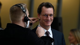 Ein Tontechniker befestigt ein Mikrofon am Gesicht von NRW-Ministerpräsident Hendrik Wüst. Der lächelt in die Kamera.
