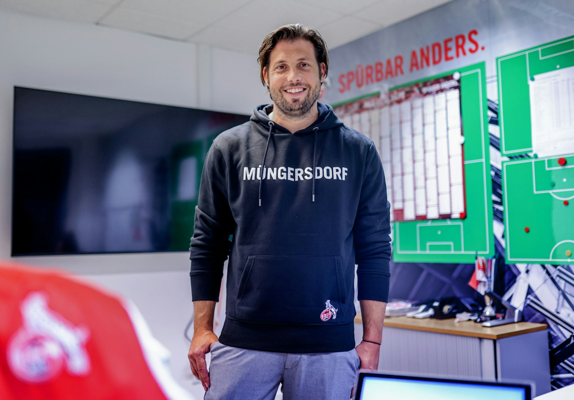 Marius Laux, Teammanager beim 1. FC Köln, steht in seinem Büro am Geißbockheim vor einem großen Jahreskalender und einem Bildschirm.