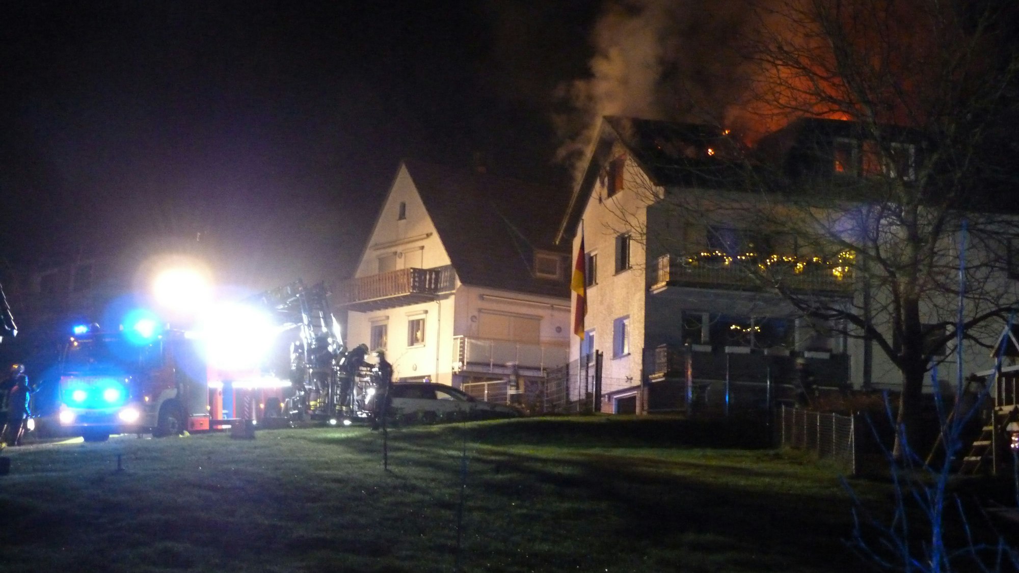 Feuerwehrwagen und Einsatzkräfte stehen auf der Straße vor dem brennenden Haus. Die Flammen sind durch das Dachfenster zu sehen.