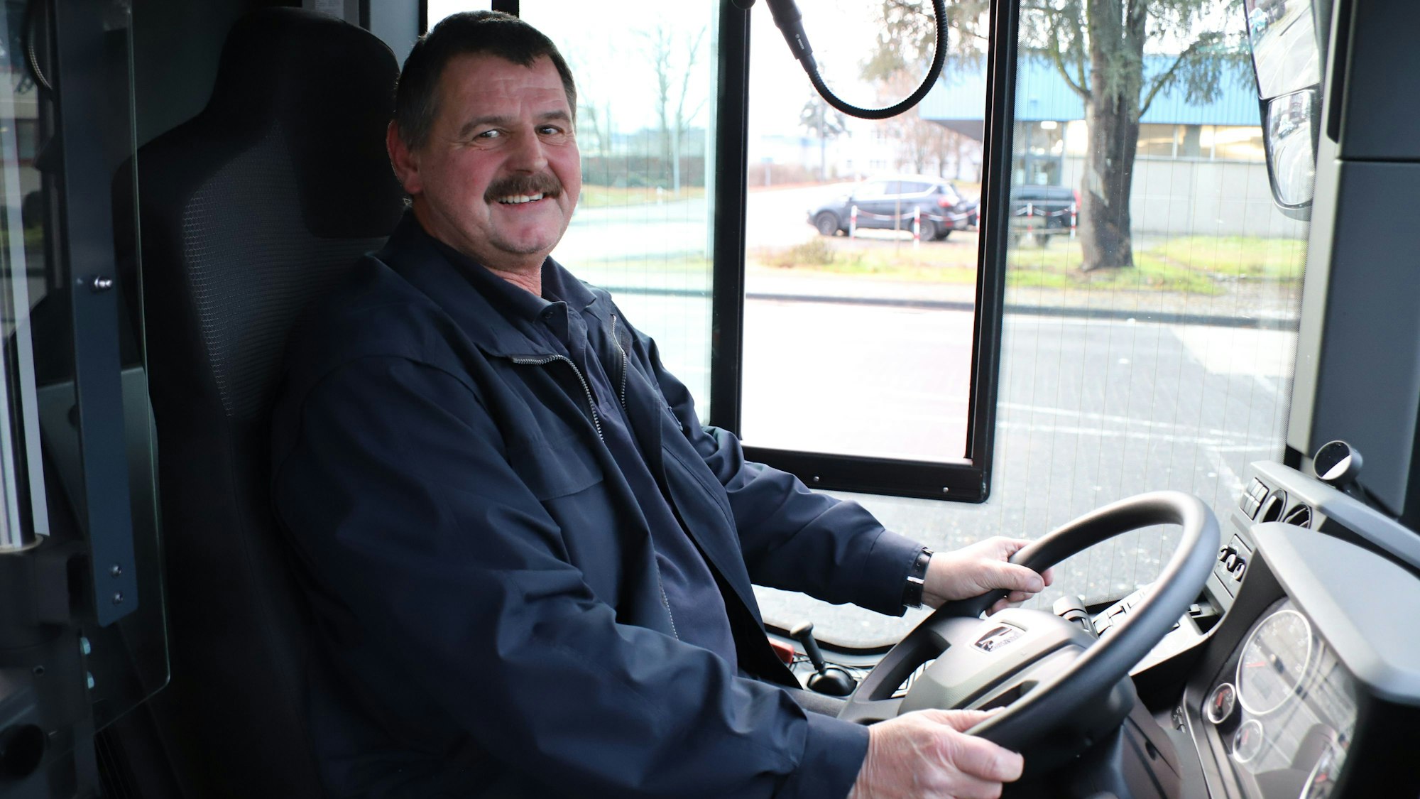 Ein Mann mit Schnurrbart sitzt am Steuer eines Busses, seine Hände halten das Lenkrad.