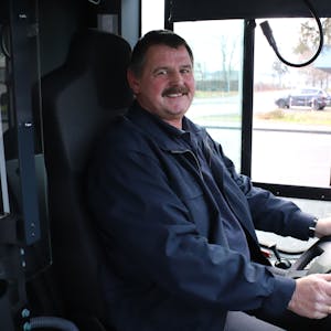 Ein Mann mit Schnurrbart sitzt am Steuer eines Busses, seine Hände halten das Lenkrad.&nbsp;