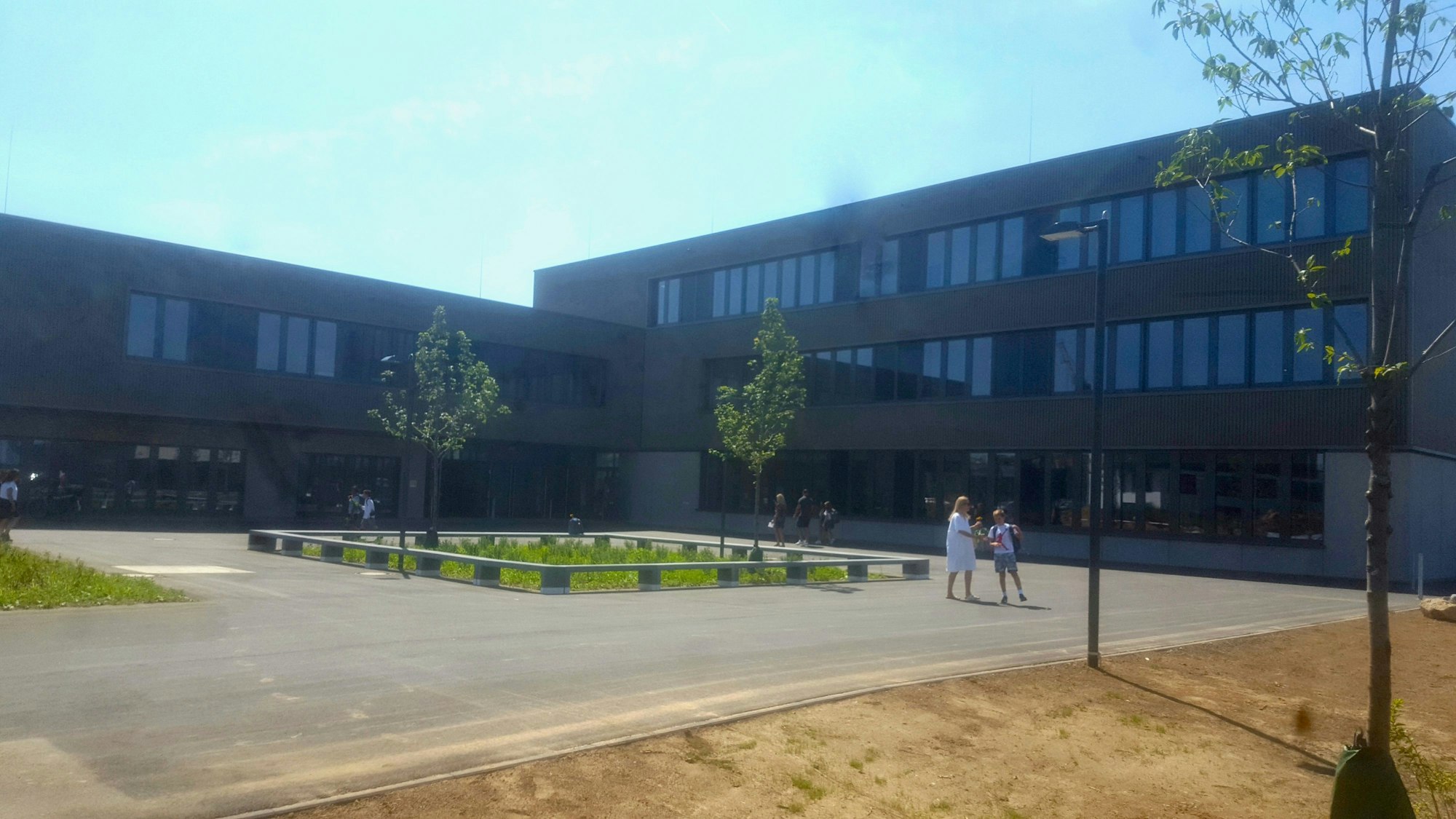 Auf dem Foto ist das neue Gymnasium ander Zusestraße in Köln zu sehen. Im Vordergrund ist eine Asphaltfläche mit Bänken und einzelnem Grün, im Hintergrund ein großer Kastenbau mit hohen Fenstern.