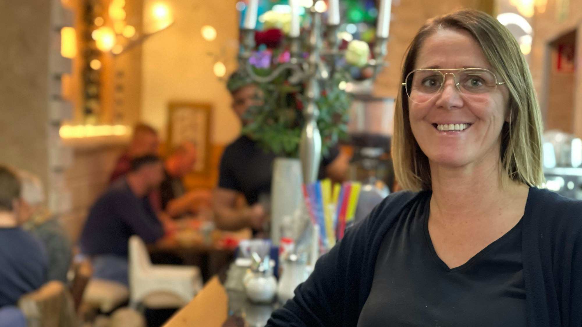 Eine Frau mit Brille, dunklem T-Shirt und Jacke lächelt. Im Hintergrund sitzen Menschen an Tischen und essen.