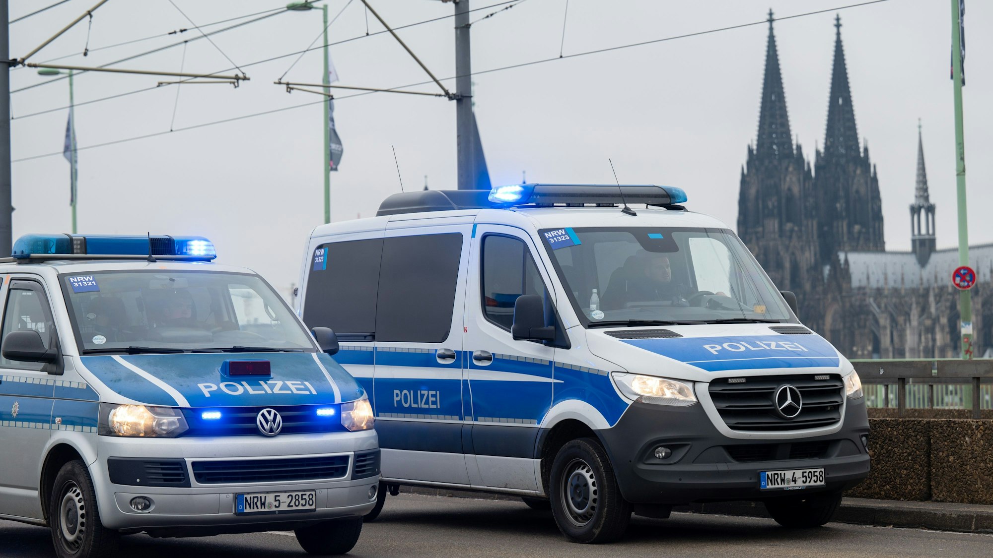 Zwei Fahrzeuge der Polizei fahren auf einer Straße, im Hintergrund ist der Kölner Dom zu sehen.