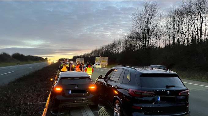 Zwei dunkle Fahrzeuge, ein Mercedes und ein BMW, stehen nach einem Zusammenstoß Blech an Blech auf der Autobahn neben der Mittelleitplanke. Menschen in Warnwesten stehen dahinter.