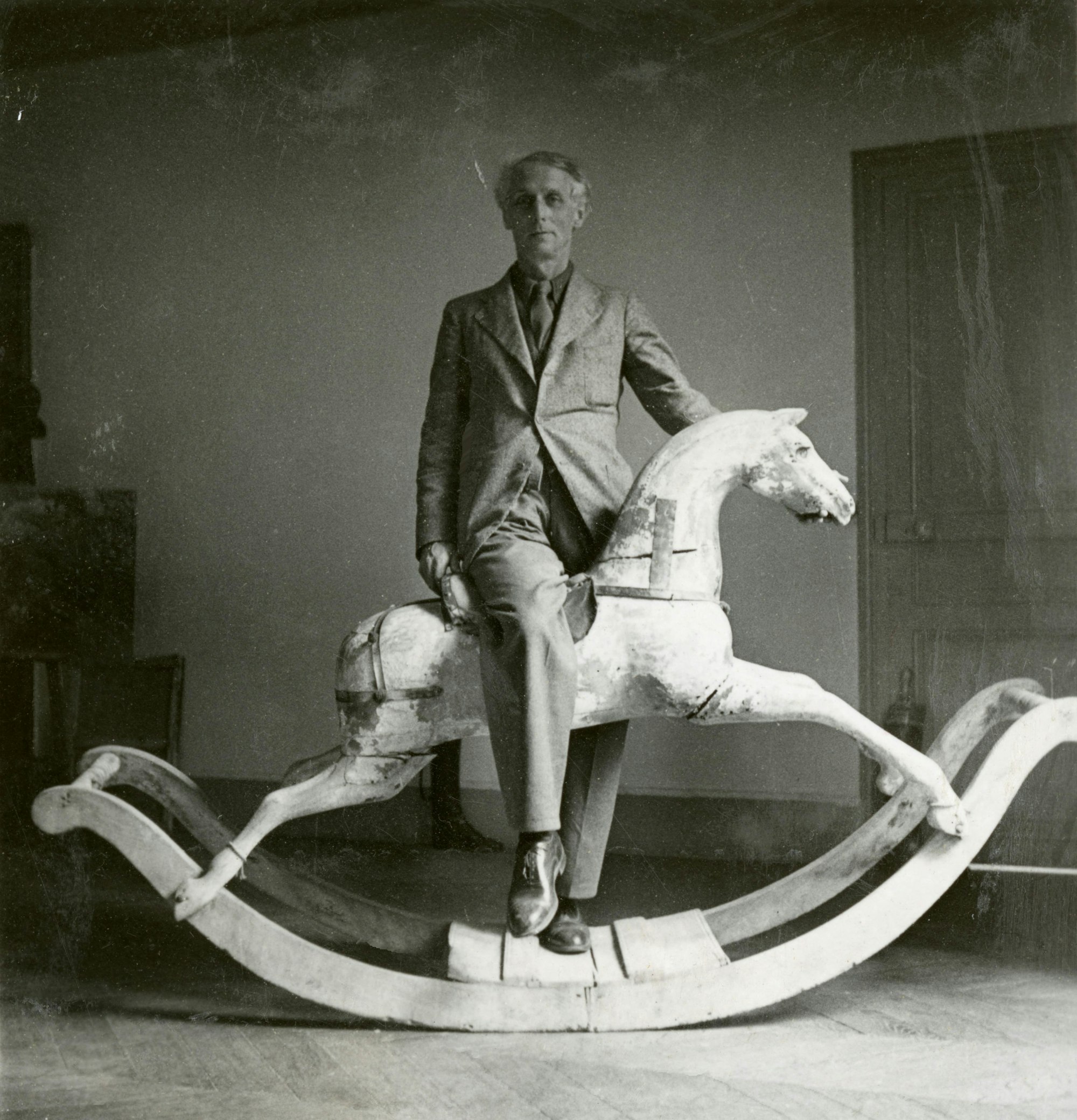 Der Surrealist Max Ernst blickt in die Kamera, ein Bein hat er quer über den Sattel eines Schaukelpferds gelegt.