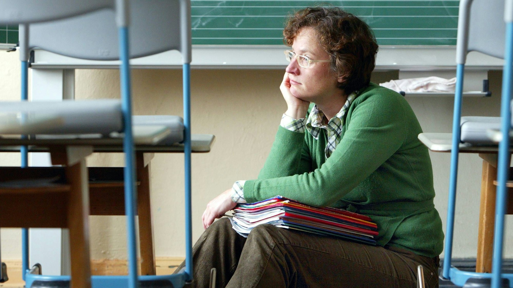 Eine Lehrerin sitzt auf dem Boden eines Klassenzimmers zwischen leeren Stühlen, hinter sich die Tafel. Auf dem Schoß hält sie einen Stapel von Heften im DIN-A4-Format. Eine Hand aufgestützt, schaut sie frustriert und traurig nach links.
