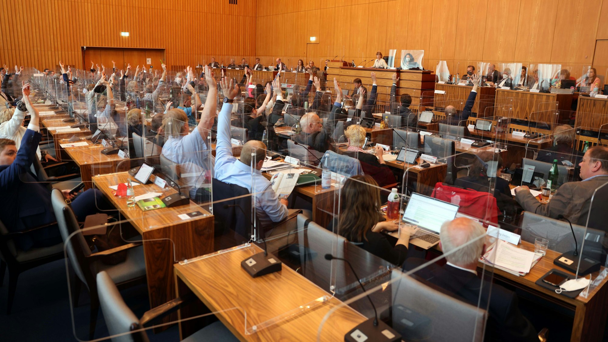 Die Mitglieder des Kölner Stadtrats sitzen an Tischen und heben die Hand für eine Abstimmung. An der Wand befindet sich die Tribüne des Präsidiums.