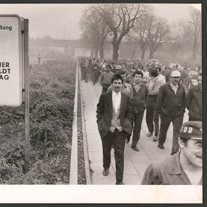 Männer gehen auf dem Schwarz-weiß-Foto in Arbeitskleidung in einem Streikmarsch am Torschild des Werksgeländes vorbei.&nbsp;