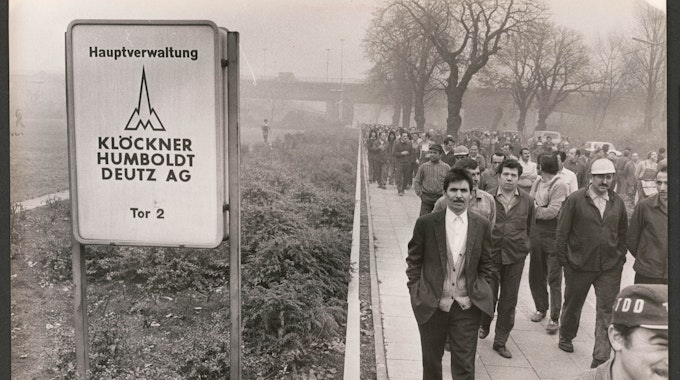 Männer gehen auf dem Schwarz-weiß-Foto in Arbeitskleidung in einem Streikmarsch am Torschild des Werksgeländes vorbei.