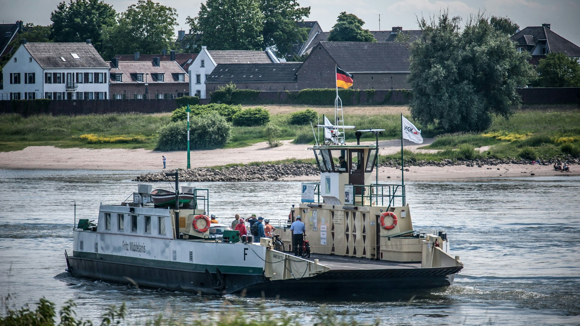 Die Rheinfähre fährt auf dem Rhein zwischen Leverkusen-Hitdorf und Köln-Langel. Ein Auto und mehrere Radfahrende sind auf dem Deck zu erkennen.