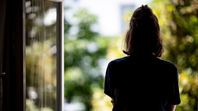 Eine Frau steht in ihrer Wohnung an einem geöffneten Fenster und blickt hinaus.