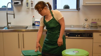 Anna Krechel steht in grüner Schürze in einer Küche neben den Mülleimern.