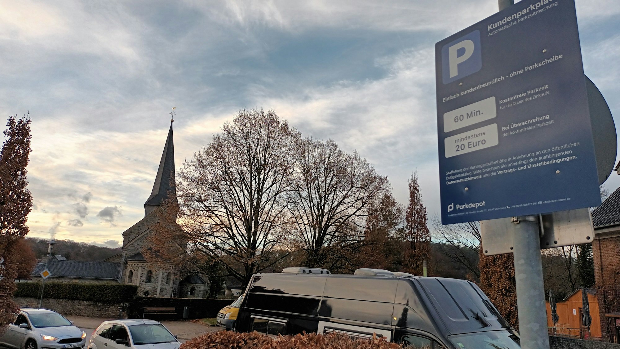Ein kleines, blaues Schild auf einem Supermarkt-Parkplatz im Lohmarer Zentrum informiert über die Höchstdauer fürs Parken und das Verwarngeld von mindestens 20 Euro. Im Hintergrund ist die katholische Kirche zu sehen.