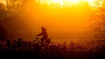 Eine Person in Mütze und dicker Jacke fährt auf einem Fahrrad durch eine Wiesenlandschaft. Die Sonne geht gerade auf.