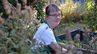 Küchenhilfe Anna sitzt in einem Garten und trägt ein weißes Shirt und eine grüne Schürze.
