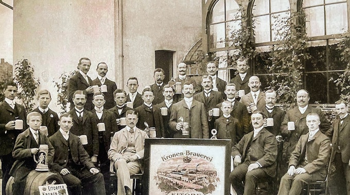 Das Bild zeigt eine Gruppe Männer mit Bierkrügen in der Hand. Sie posieren vor der Brauerei für ein Foto.