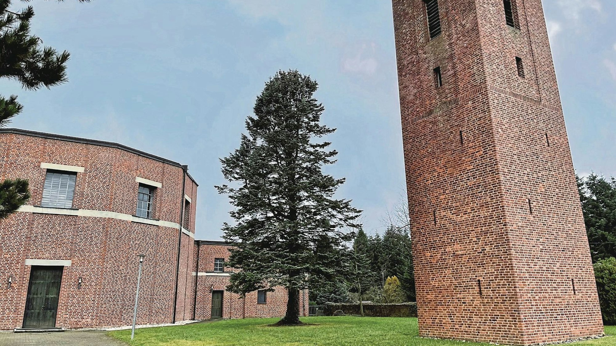 Rechts steht ein hoher, viereckiger Backsteinturm, links ein Rundbau ebenfalls aus Backsteinen. Das Gebäudeensemble liegt in einem kleinen Park, in dem ein hoher Nadelbaum steht.