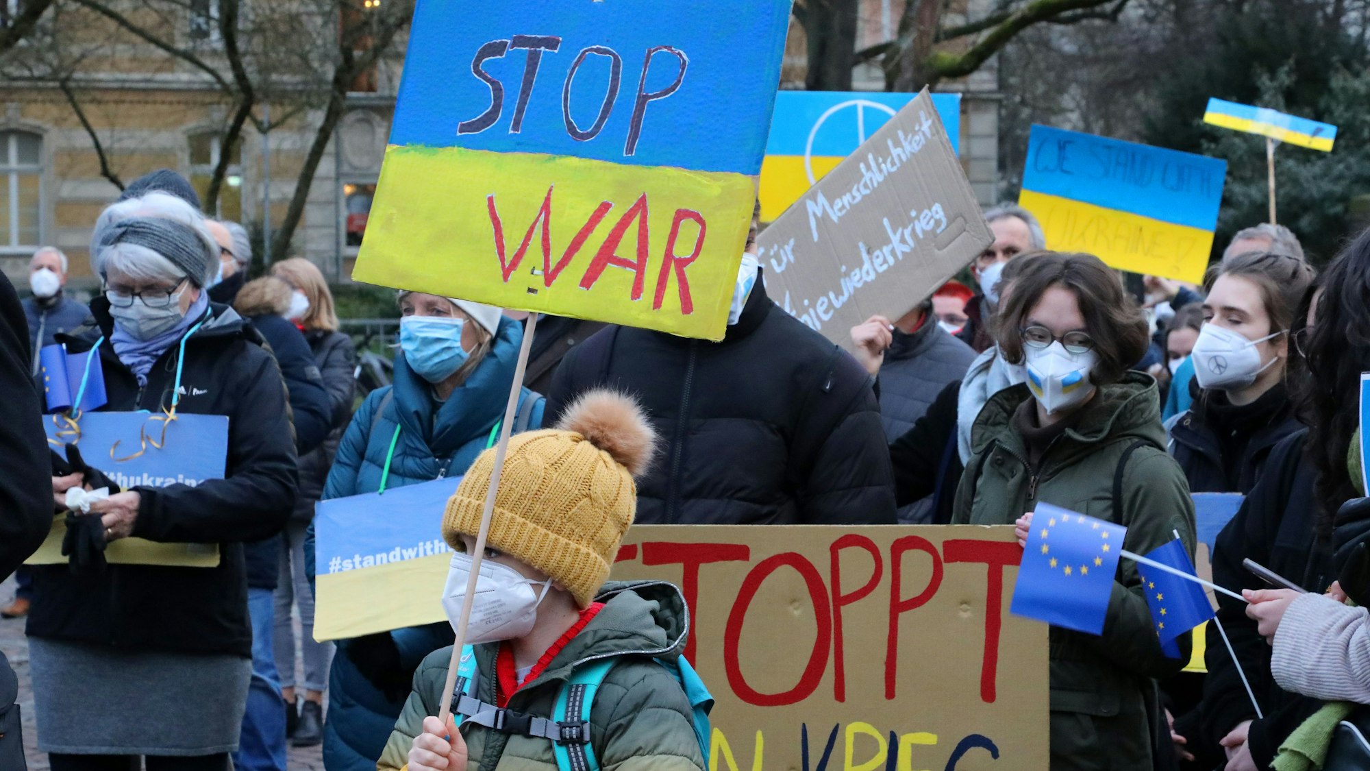 Menschen mit Plakaten auf den "Stop War" und "Stoppt den Krieg" zu lesen ist, stehen bei einer Demonstration gegen den Krieg in der Ukraine auf dem Konrad-Adenauer-Platz in Bergisch Gladbach.