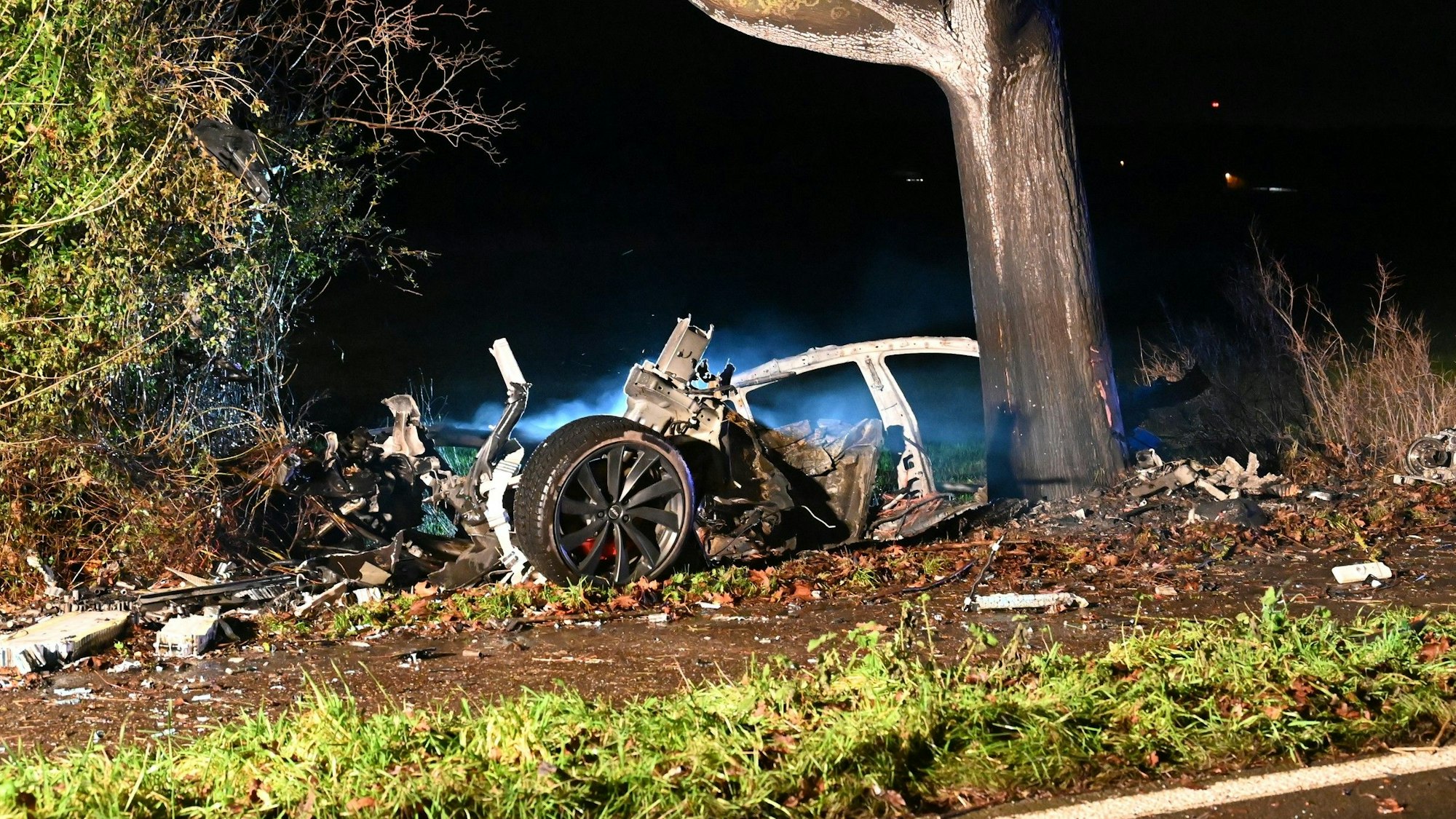 Zwischen Bergheim und Pulheim ist ein Tesla am 31.12.2022 auf der L213 ausgebrannt. Überreste des Wagens liegen am Straßenrand, daneben steht ein Baum.
